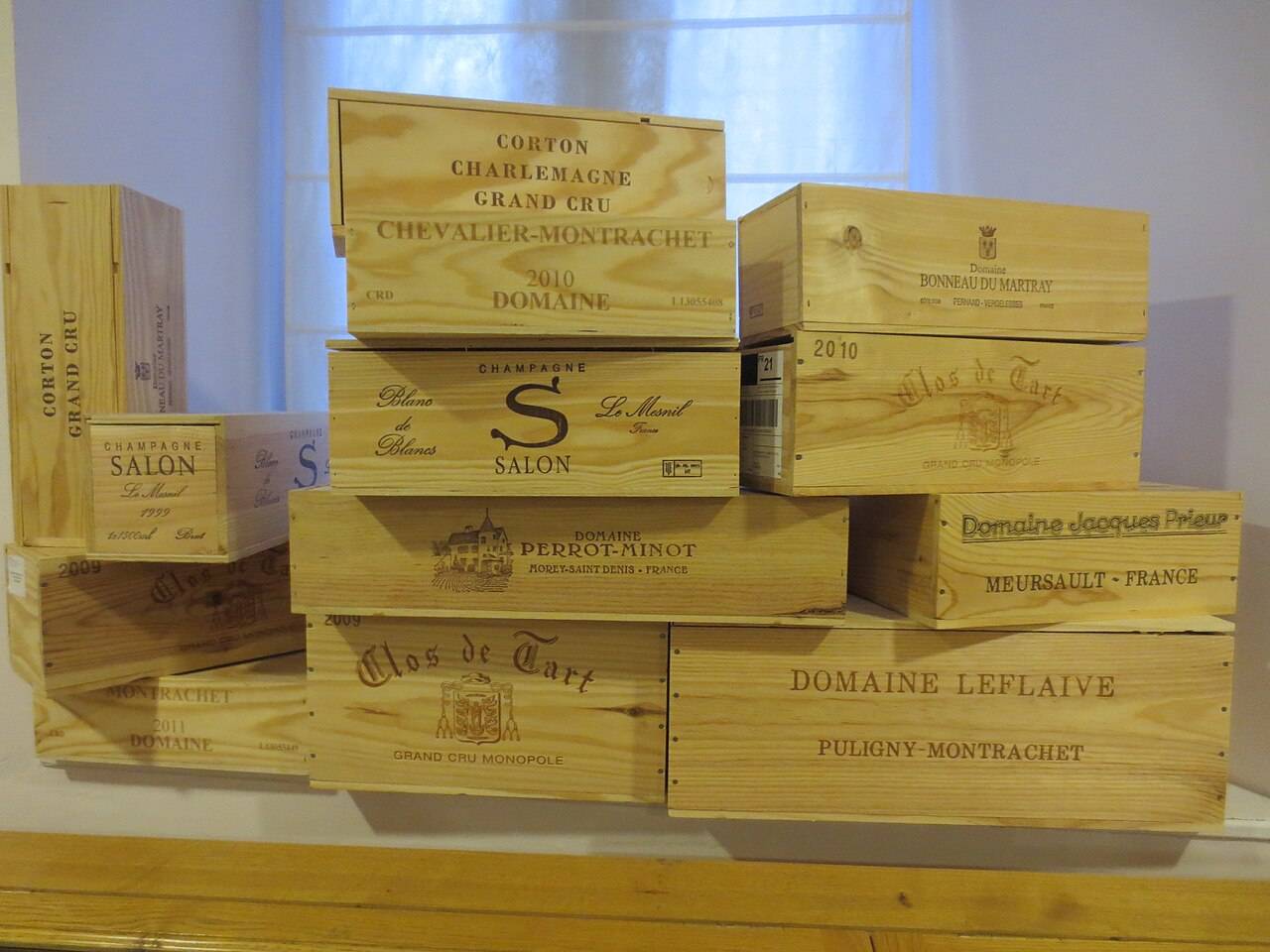 Des caisses en bois empilées aux noms de grands vins : Clos de Tart, Domaine Leflaive, Corton-Charlemagne, Chevalier-Montrachet et champagne Salon