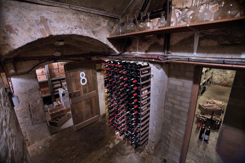 Une cave voûtée en brique, avec un casier métallique garni de bouteilles couchées et une porte numérotée