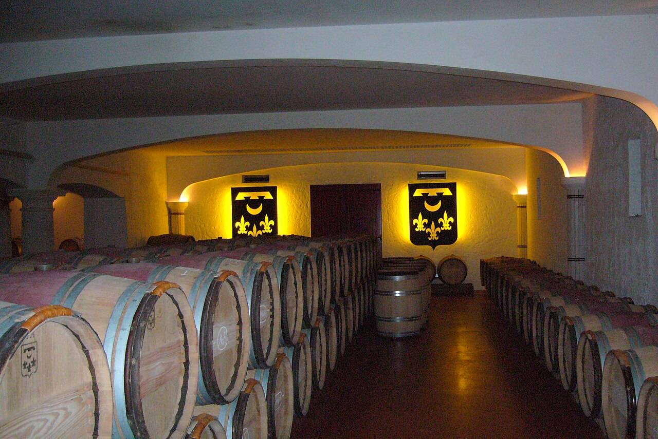 Des rangées de barriques sous les voûtes éclairées d’un chai du Château Smith Haut Lafitte, devant deux blasons aux fleurs de lys