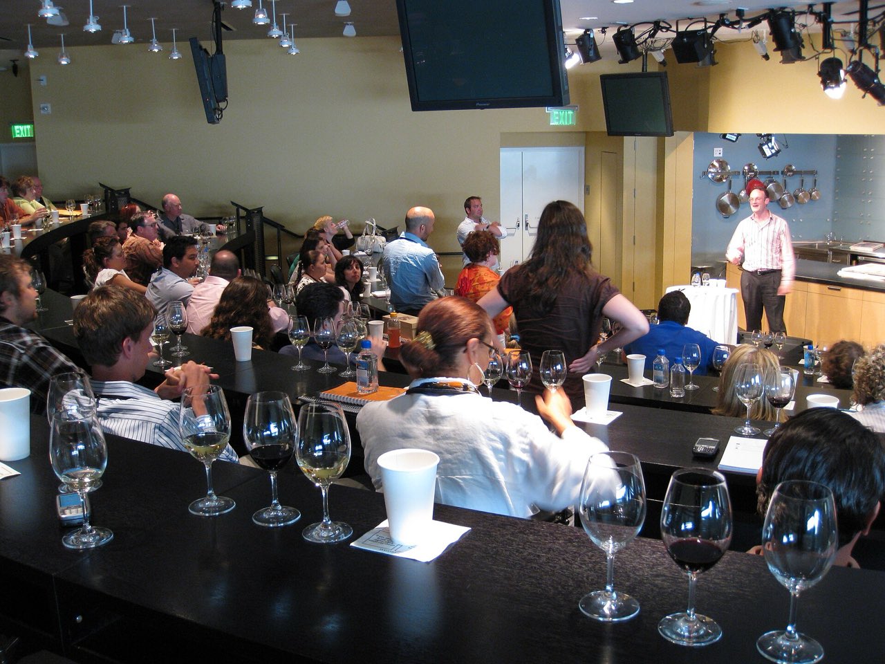 Une salle de cours pleine de participants assis derrière des rangées de verres à vin, face à un animateur