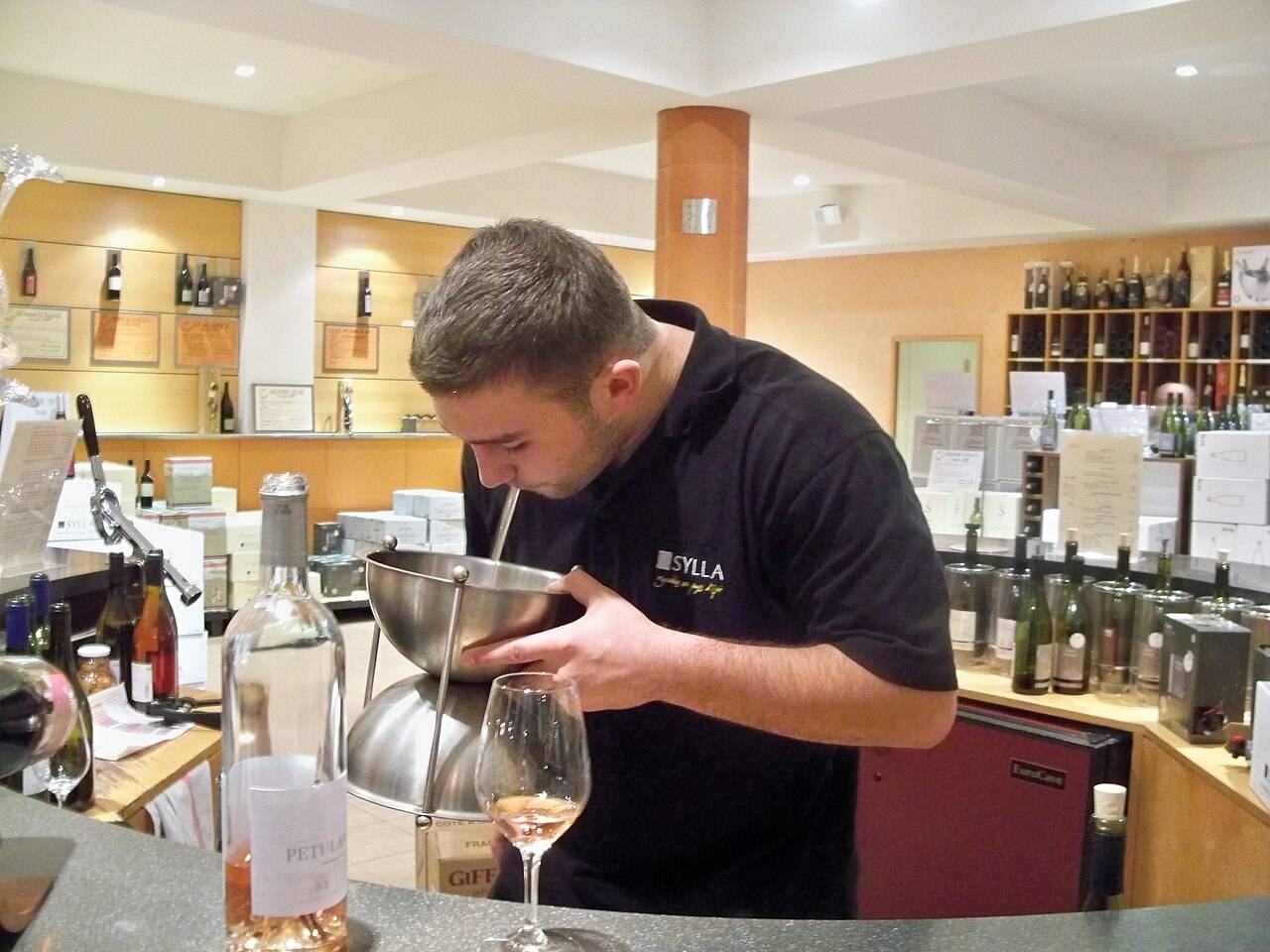 Un homme recrache le vin dans un crachoir en métal, au comptoir d’un caveau, à côté d’un verre et d’une bouteille de rosé