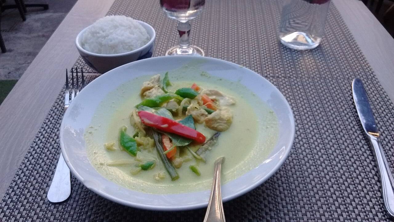 Une assiette de curry vert thaï au lait de coco, avec un bol de riz, un verre de vin et une carafe d’eau, dans un restaurant parisien