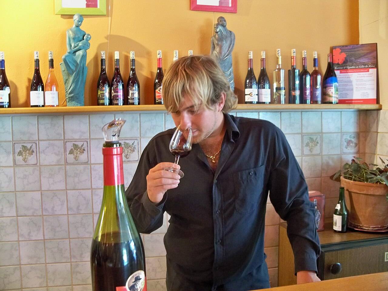 Un homme hume un verre de vin rouge au comptoir d’un caveau de dégustation, devant une étagère de bouteilles