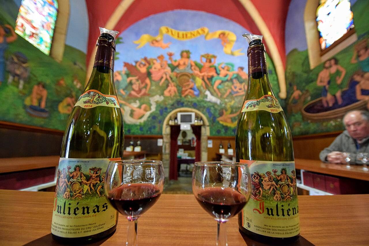 Deux bouteilles de Juliénas et deux verres de vin rouge sur un comptoir, devant des fresques bachiques et des vitraux