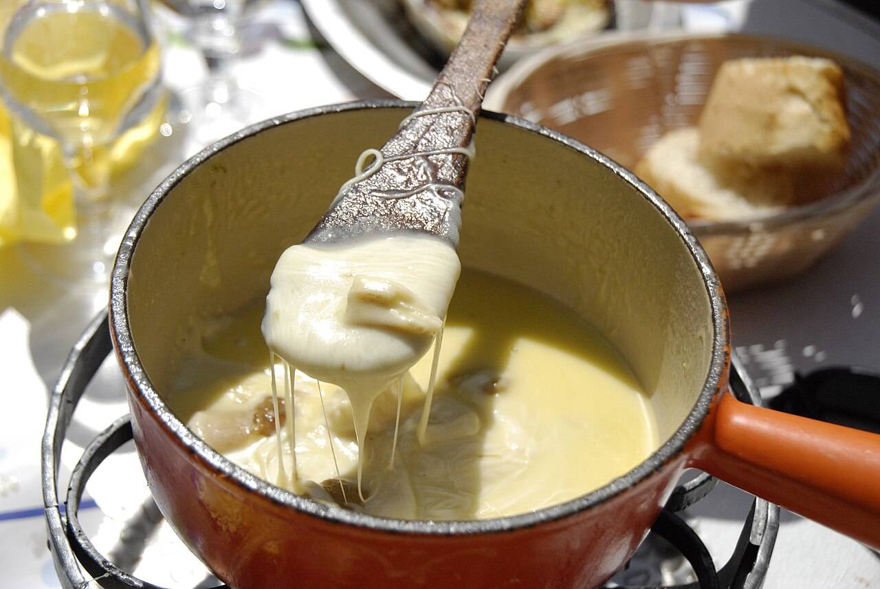 Un caquelon de fondue savoyarde au Beaufort et aux champignons, d’où l’on tire un morceau de pain nappé de fromage, avec un verre de vin blanc à l’arrière-plan