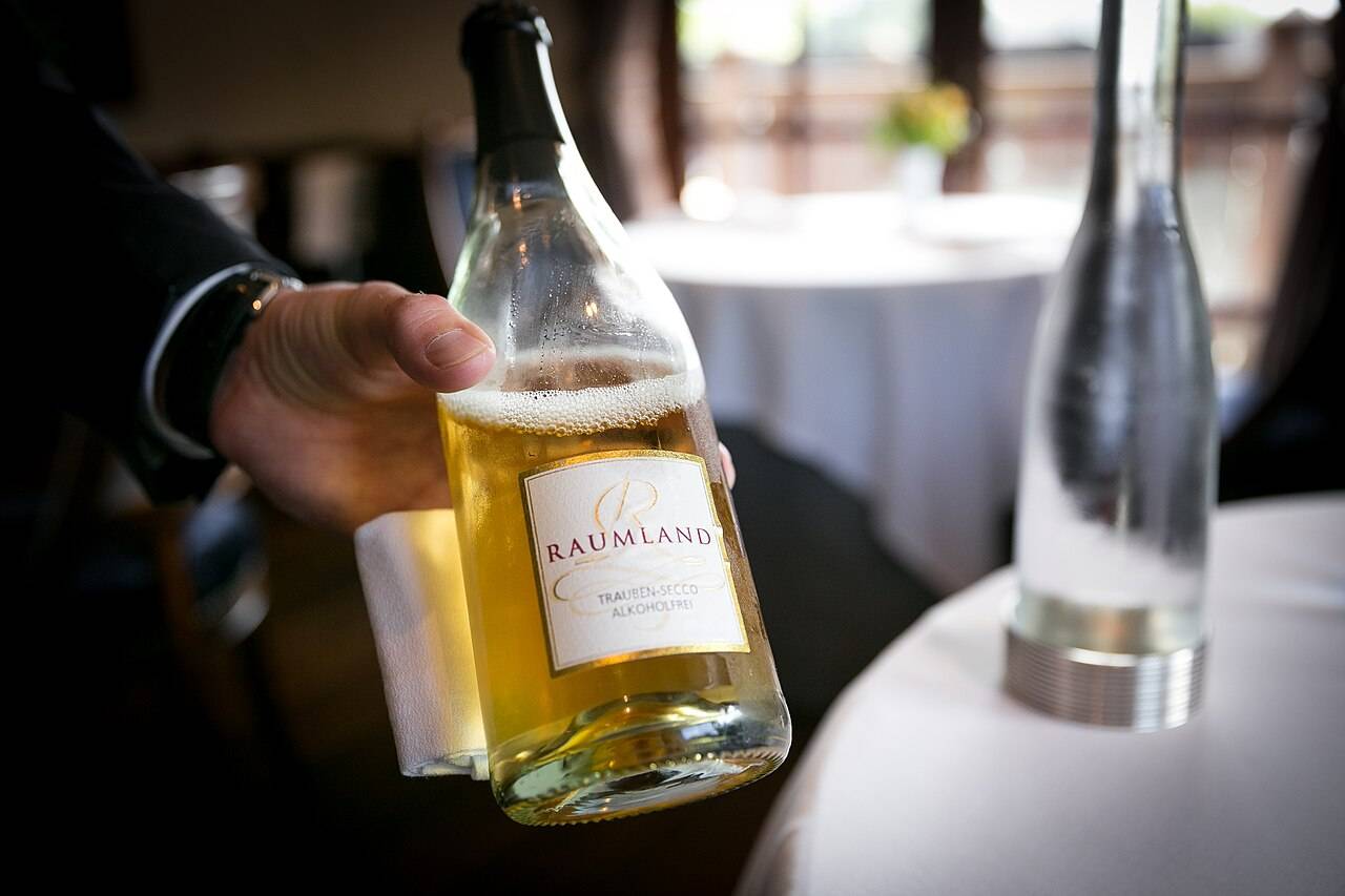 Un serveur présente une bouteille entamée de Raumland Trauben-Secco sans alcool, un pétillant de raisin allemand, à côté d’une carafe d’eau
