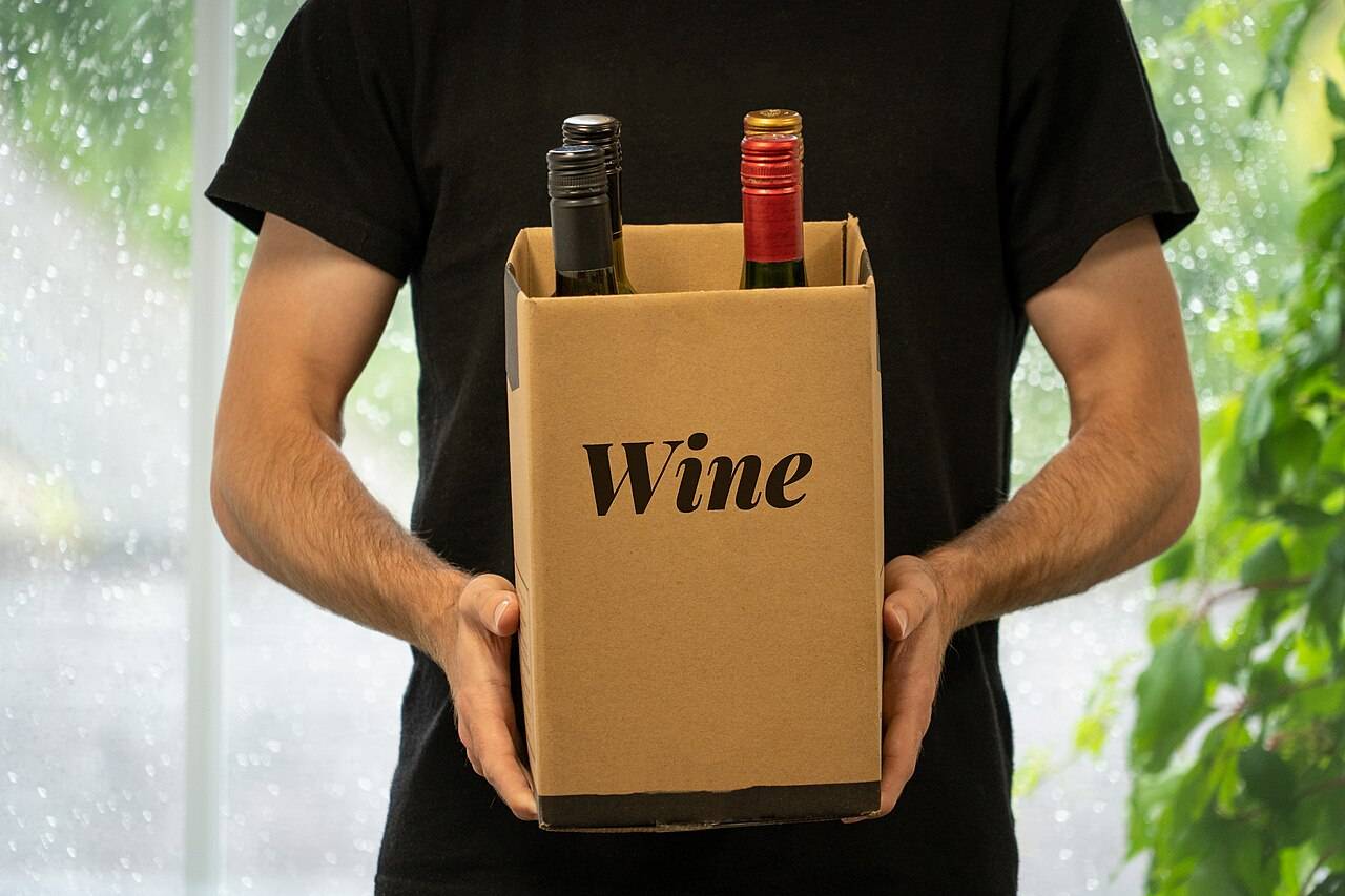 Un homme tient à deux mains un carton marqué «&nbsp;Wine&nbsp;», d’où dépassent les goulots de quatre bouteilles