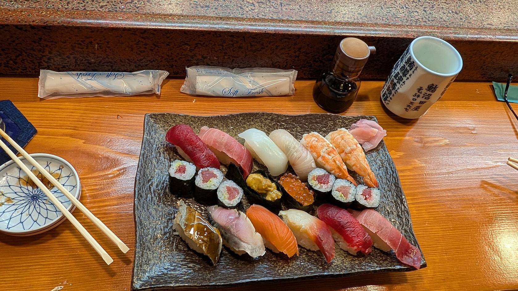 Un plateau de nigiris et de makis sur le comptoir d’un bar à sushis, avec des baguettes, de la sauce soja et une tasse de thé