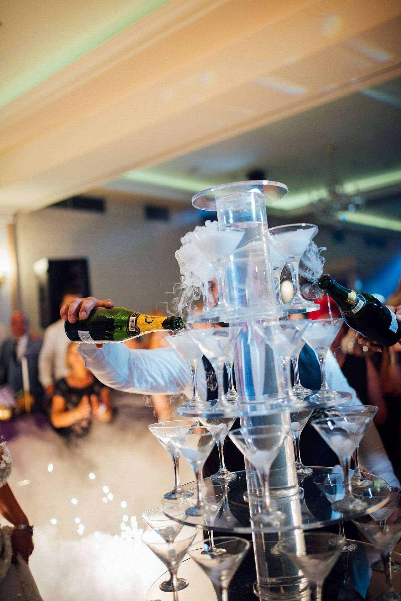 Une pyramide de coupes que l’on remplit d’un vin effervescent pendant une réception, dans un nuage de fumée, avec des invités autour