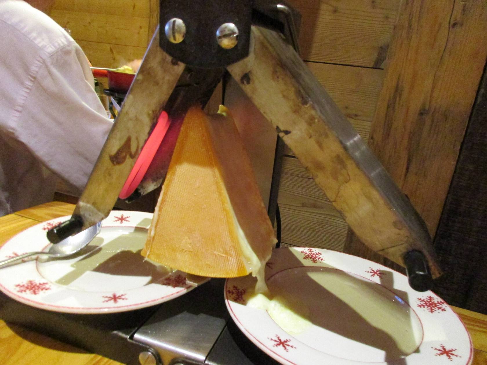 Un quartier de fromage à raclette fixé dans un appareil en bois, dont le fromage fondu est raclé sur des assiettes