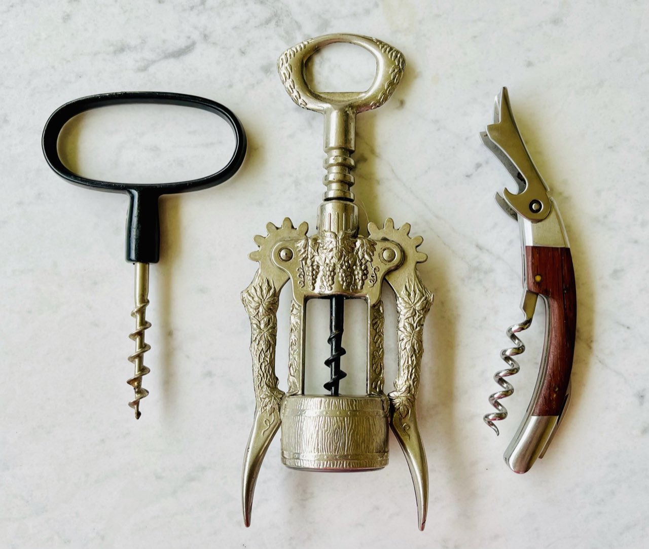 Trois tire-bouchons posés sur du marbre : un modèle à poignée, un modèle à ailettes et un tire-bouchon de sommelier